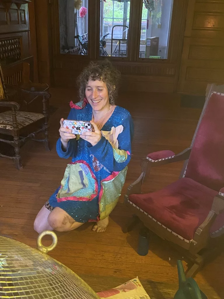 A photo of vilde, kneeling on the floor taking a picture of the person who's taking this picture. She's grinning with delight, her curly black and chrome hair is still wet from gardening in the rain, and she's wearing a blue sparkly, slouchy dress with green and pink lily pads and beetles on it.