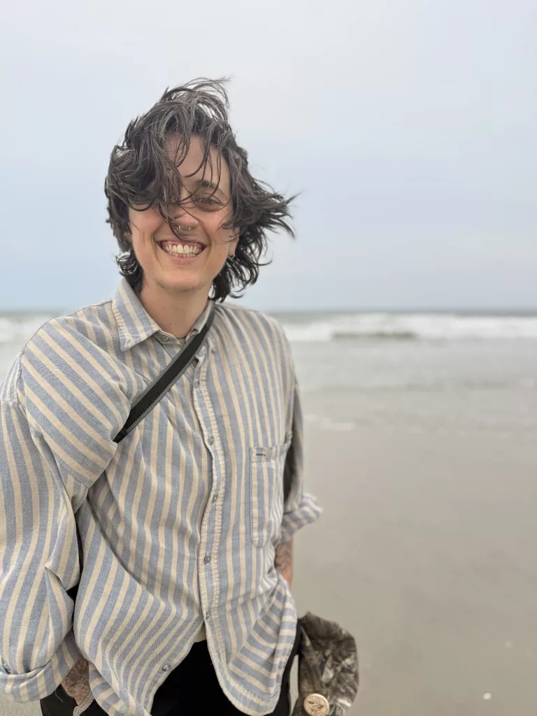 ced is on the beach on a gray day, an earnest, youthful smile on their face, soft eyes and big grin, hair blowing everywhere, while blurred waves break in the distance